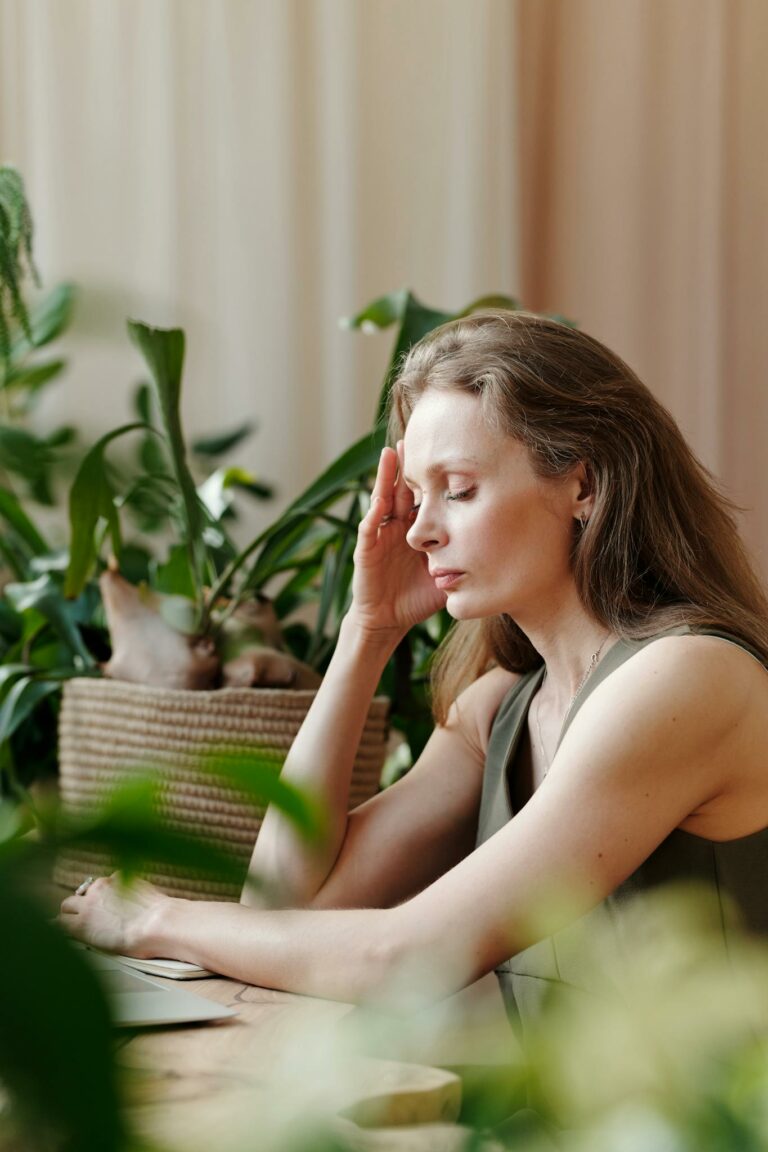Adult woman sitting at home surrounded by plants, touching her temple with eyes closed, signifying a headache.